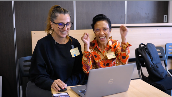 Two women are sitting at a table, smiling and using a laptop. One of the women is wearing glasses.

Contenu généré par l’IA